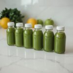 Green juice in glass bottles with celery, lemon, and ginger on white table